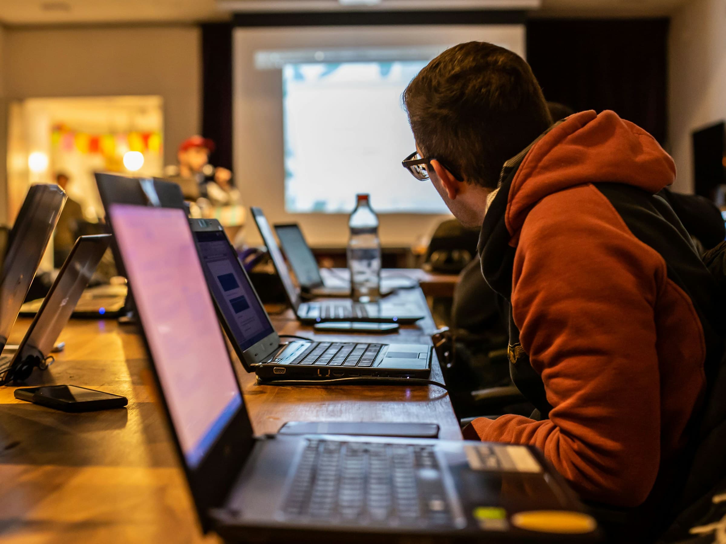 Deelnemer van achteren in een workshopzaal met een rij laptops en een projectiescherm